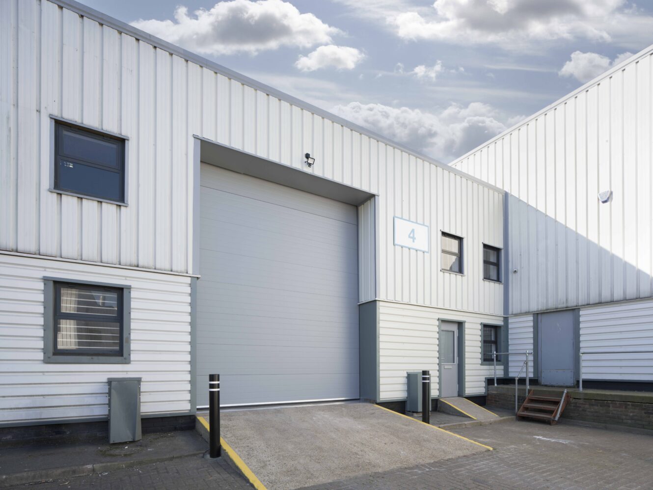 A modern industrial warehouse with a large closed roller shutter, several windows, and a number 4 sign under partly cloudy skies.