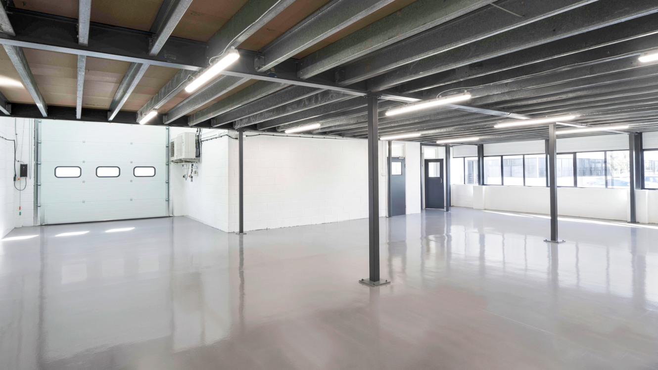 Empty industrial warehouse space featuring a polished concrete floor, exposed ceiling beams, a large garage door, and wall of windows.