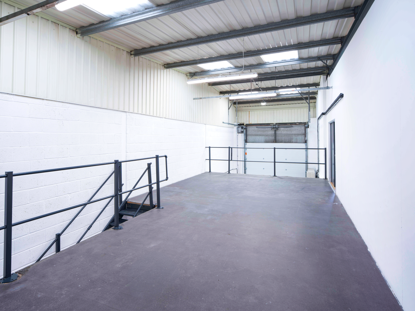 Interior view of an empty industrial mezzanine with metal railings, concrete floor, white walls, and a large roller shutter at the end.