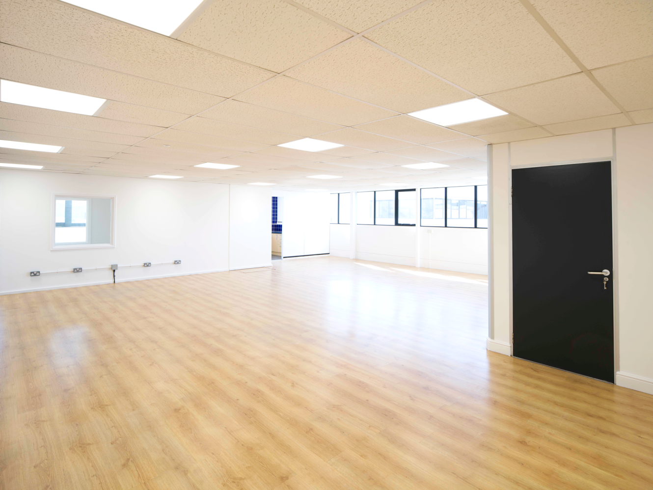 Empty office with light wood flooring, white walls, large windows, a closed black door, even ceiling lights, and natural light from the right.