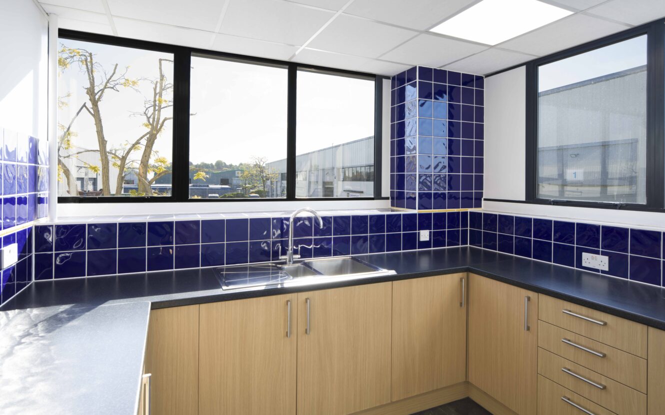 Modern kitchen featuring blue tile splashback, wooden cupboards, black worktop, stainless steel sink, and large sunlit windows.