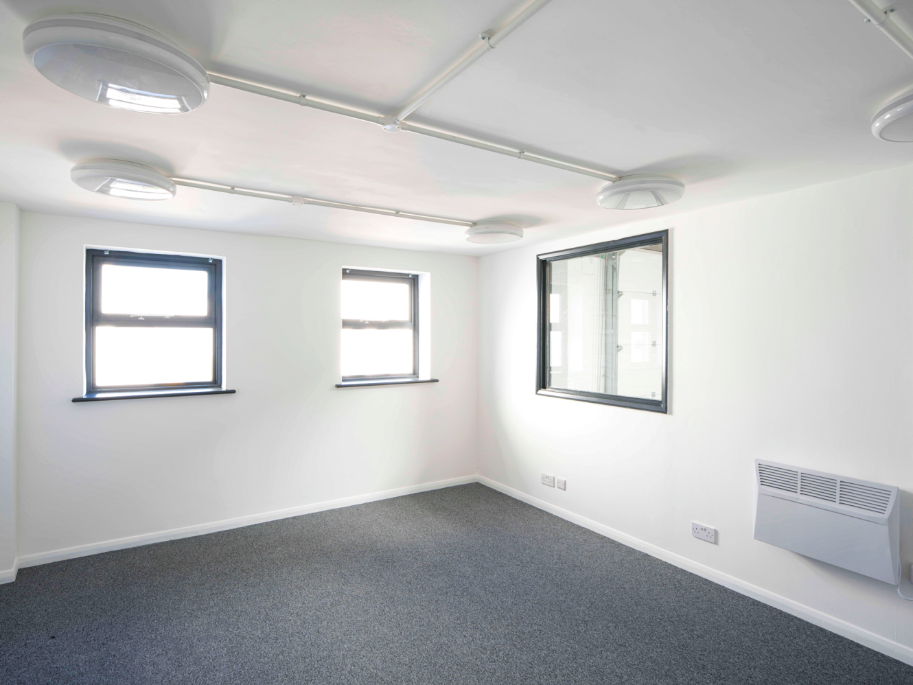 Empty, unfurnished room with white walls, grey carpet, three windows, wall-mounted heater, internal window, and exposed ceiling fittings.