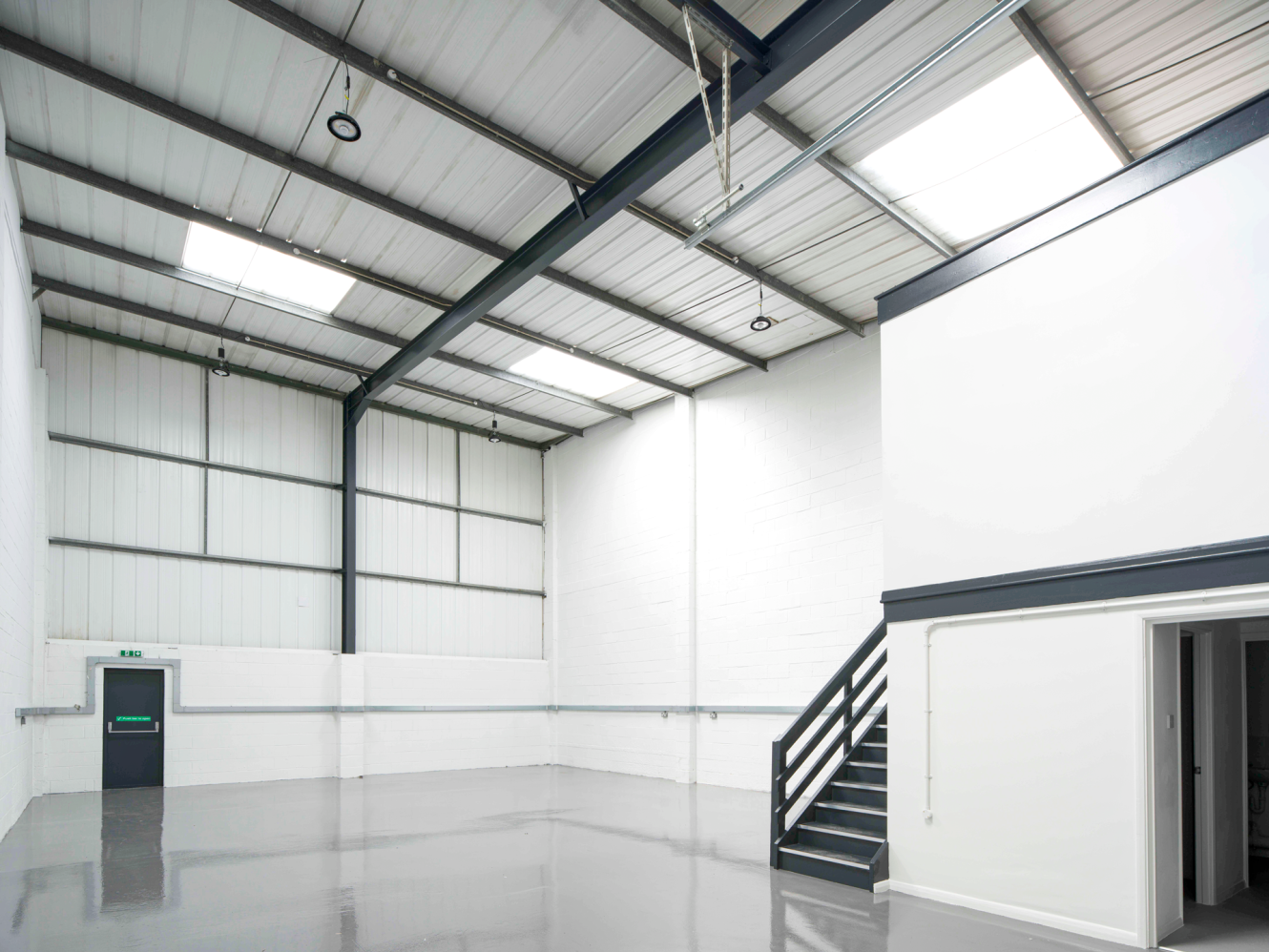 Empty industrial warehouse with high ceilings, skylights, white walls, grey floor, mezzanine staircase, and green exit door.