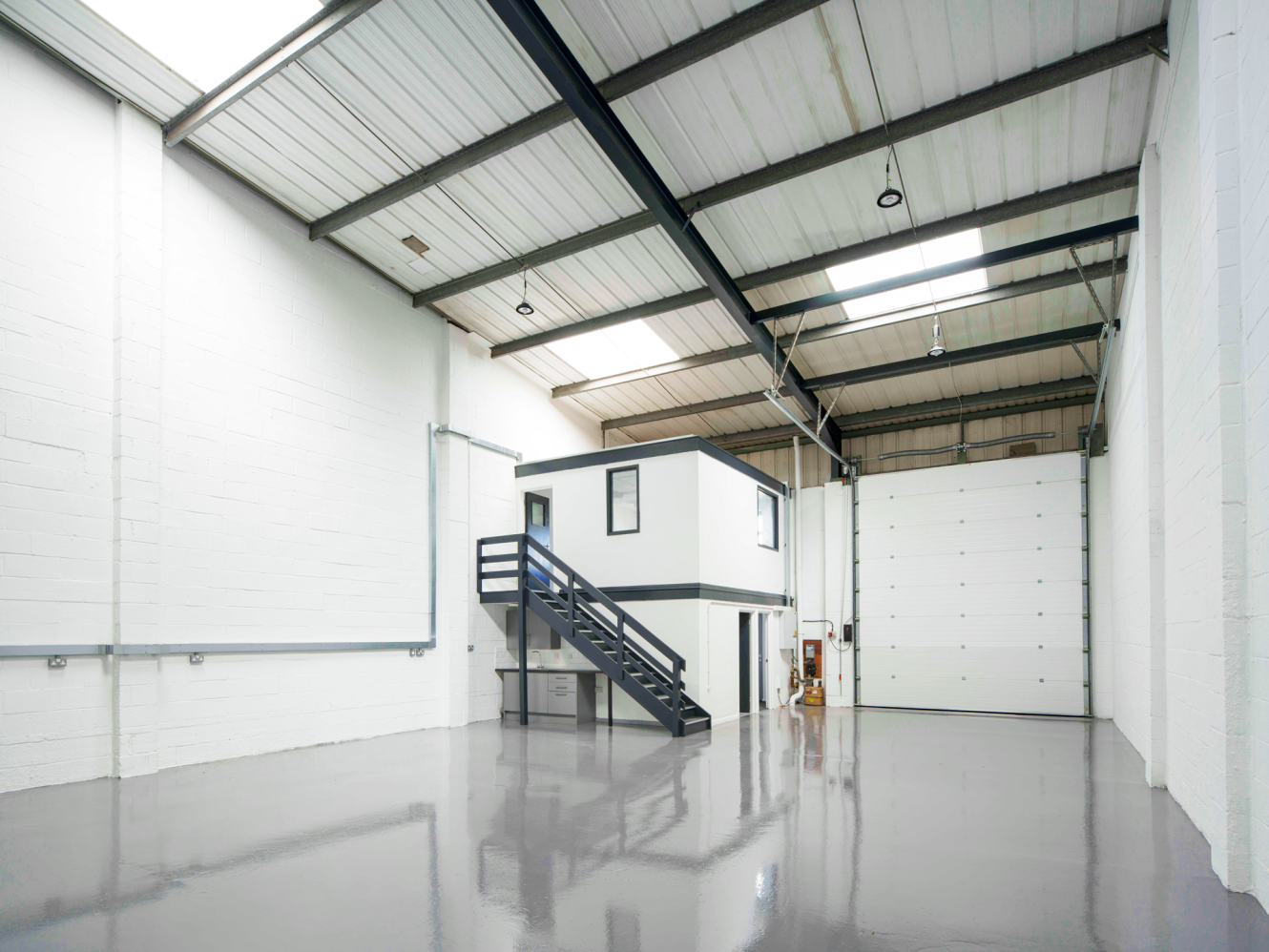 An empty industrial warehouse with white walls, a grey polished floor, mezzanine office, metal stairs, and large roller shutter door.