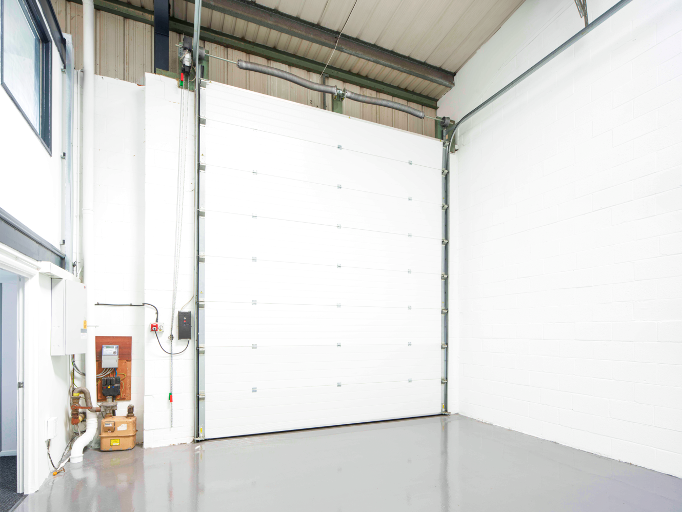 A large white industrial garage door is fitted in the corner of a clean, empty warehouse with white walls and a grey floor.