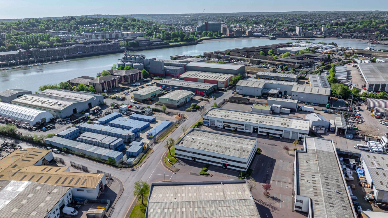 Aerial view of an industrial estate with warehouses and buildings by a river, city and greenery in the background under clear skies.