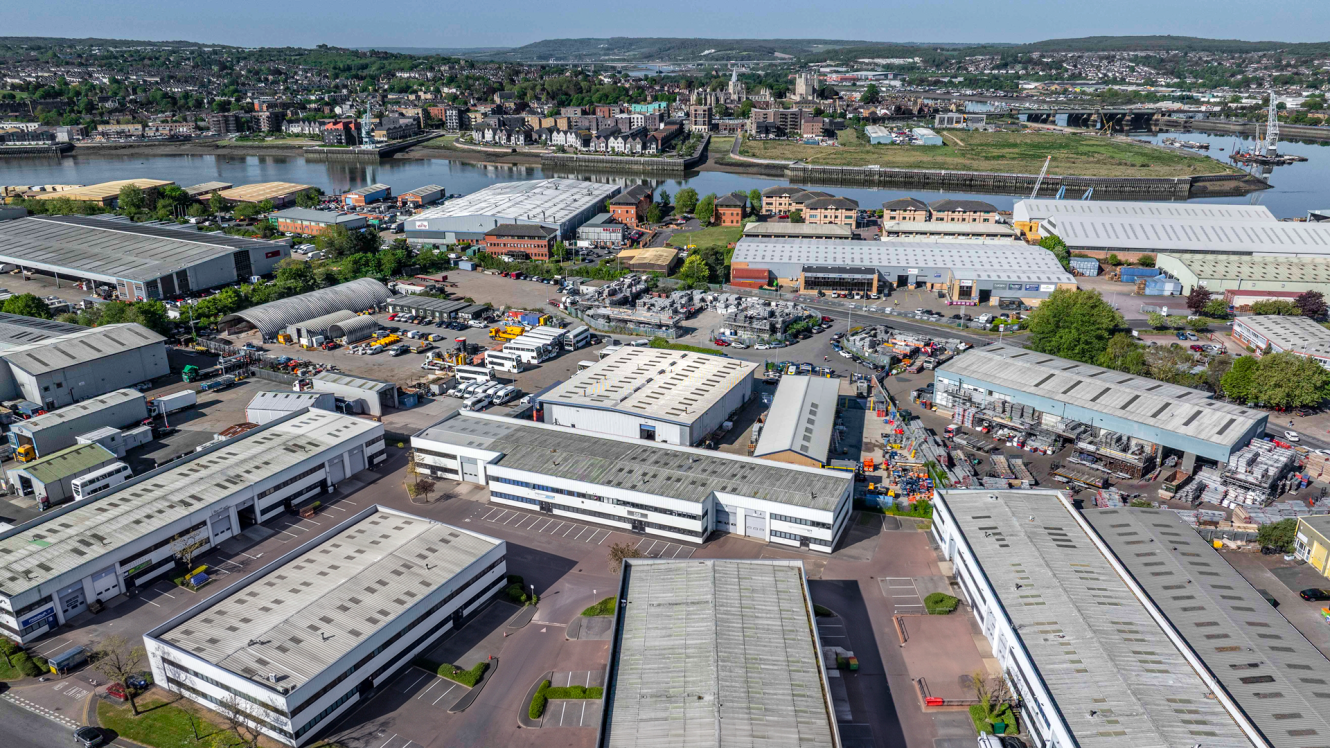 Aerial view of an industrial estate featuring warehouses, factories, car parks, and a river with a town in the background.
