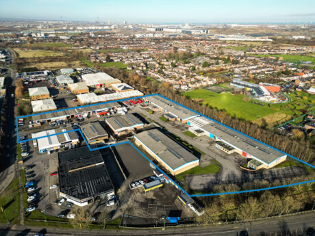 Aerial view of an industrial estate outlined in blue among homes, green areas, and roads, with a town and more industry beyond.
