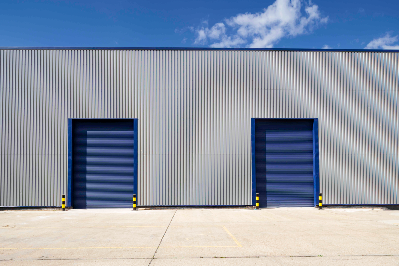 A grey industrial warehouse with two large blue roller doors, yellow and black safety bollards, and a clear sky overhead.