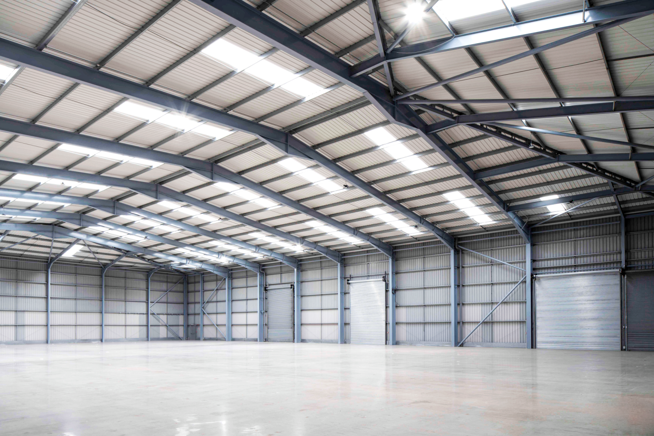 Large, empty warehouse with metal beams, high ceiling, polished concrete floor, closed roller doors, and bright skylight illumination.