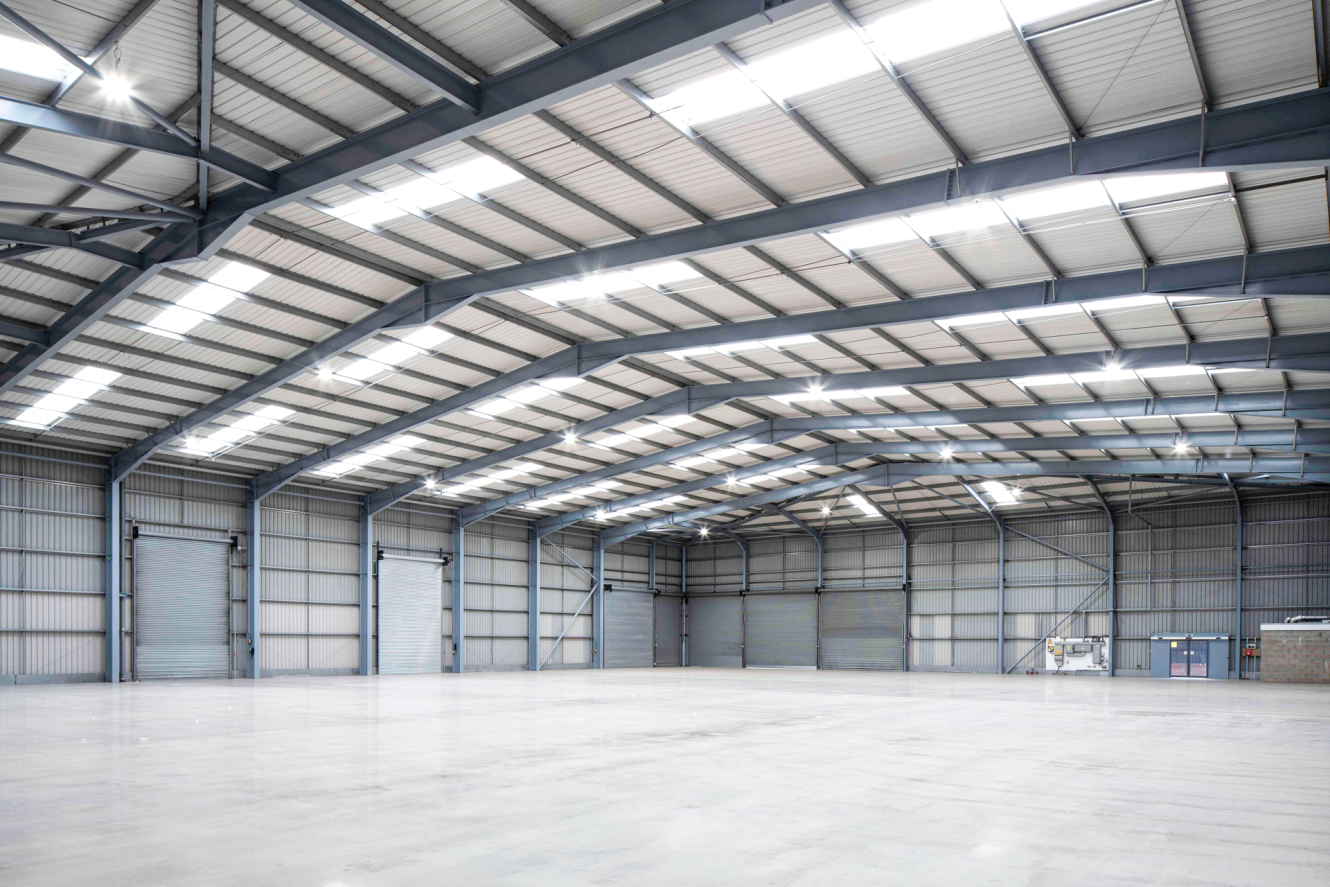 Large, empty warehouse with metal beams, high ceilings, bright overhead lighting, wide open floor space, and closed shutter doors.