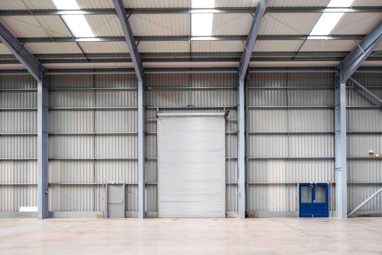 Interior of an empty industrial warehouse featuring a large closed metal roller shutter, concrete floor, and exposed steel beams.