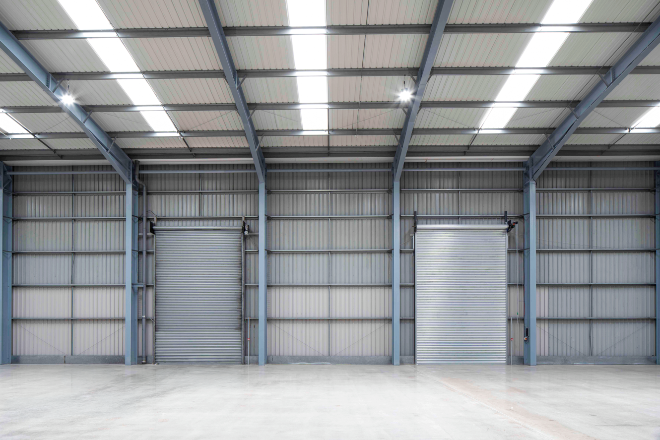 Interior of an empty warehouse with metal walls, concrete floor, and two large closed roller shutters under skylights in the ceiling.