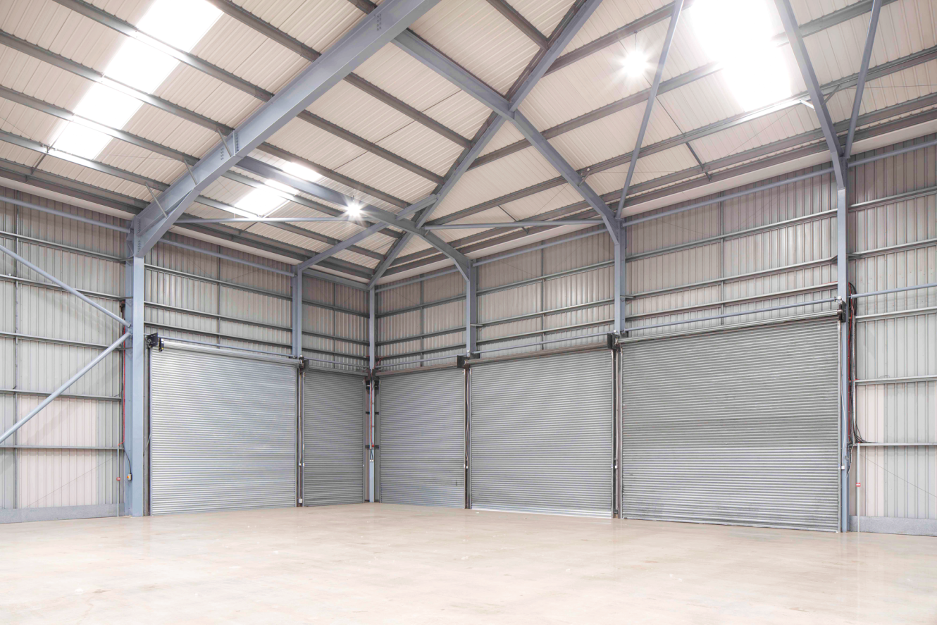 Spacious, empty warehouse with high ceilings, exposed steel beams, large overhead lights, and several closed metal roller shutters.