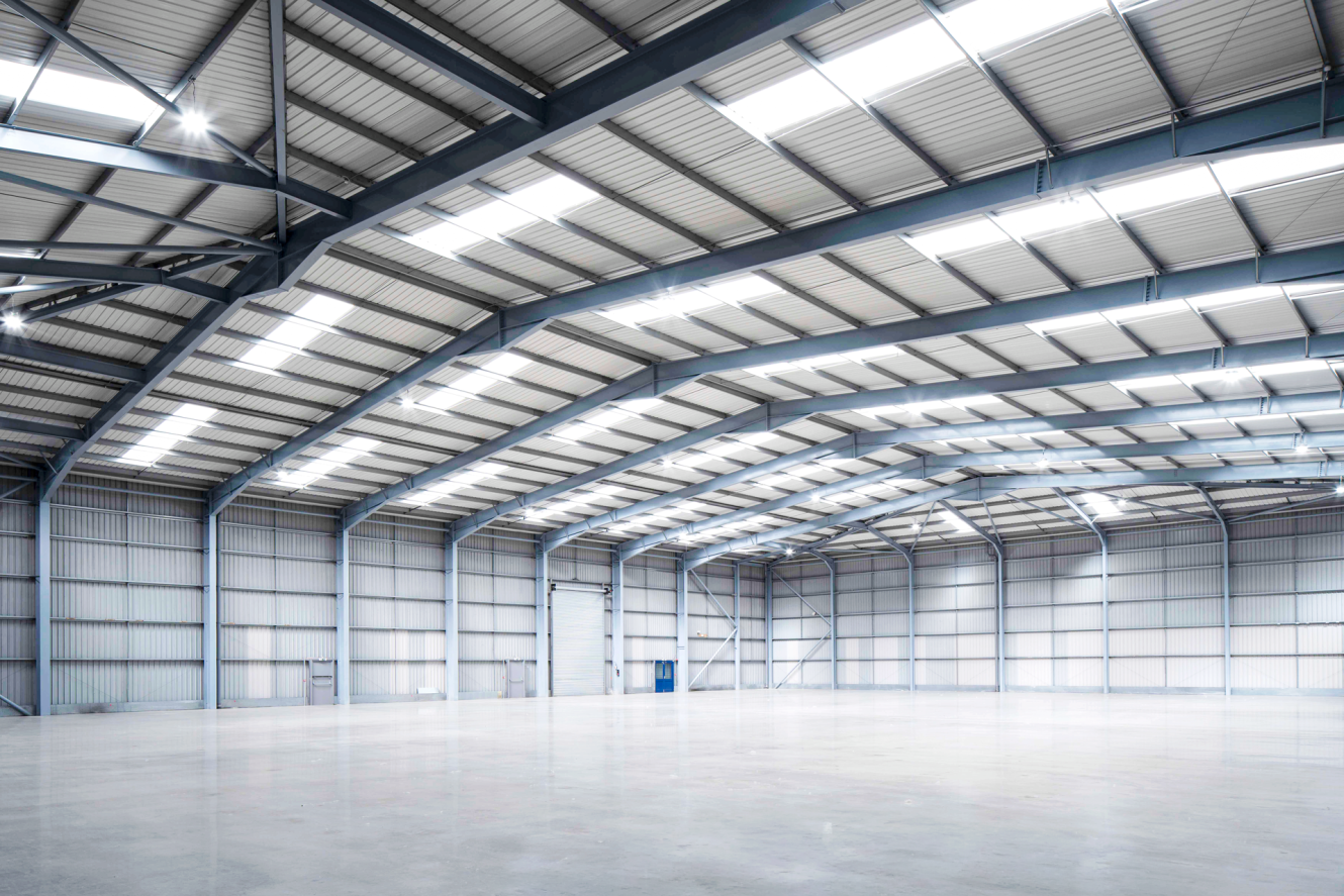 Interior of a spacious, empty warehouse featuring high ceilings, metal beams, polished concrete floors, and mixed natural and artificial light.