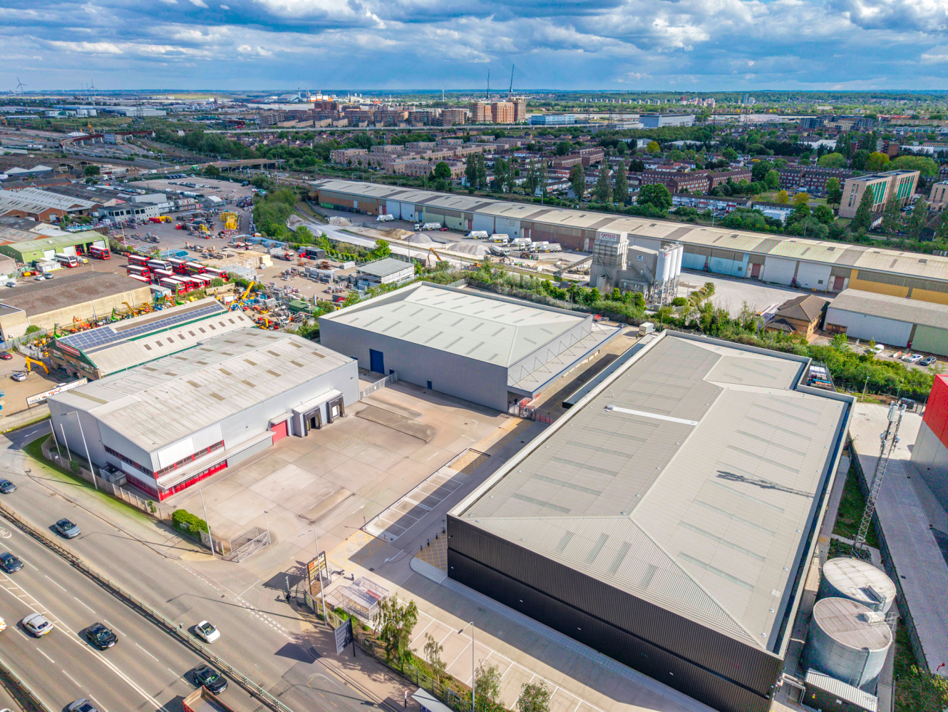 Aerial view of warehouses and factories by a main road, with residential and commercial buildings behind, under partly cloudy sky.