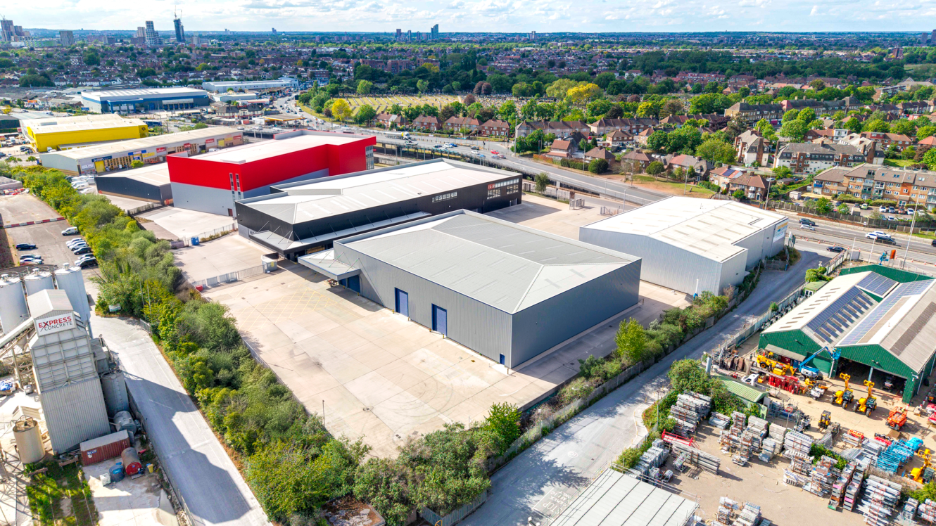 Aerial view of an industrial estate with large warehouses, red and grey exteriors, roads, trees, and nearby residential buildings.