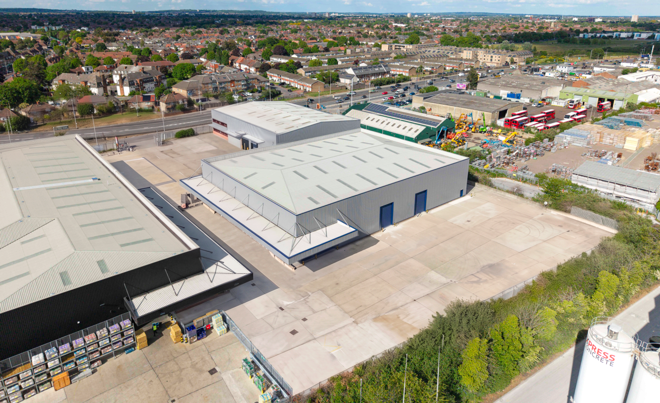 Aerial view of an industrial warehouse complex with large paved yards, next to a suburban area and various commercial buildings.