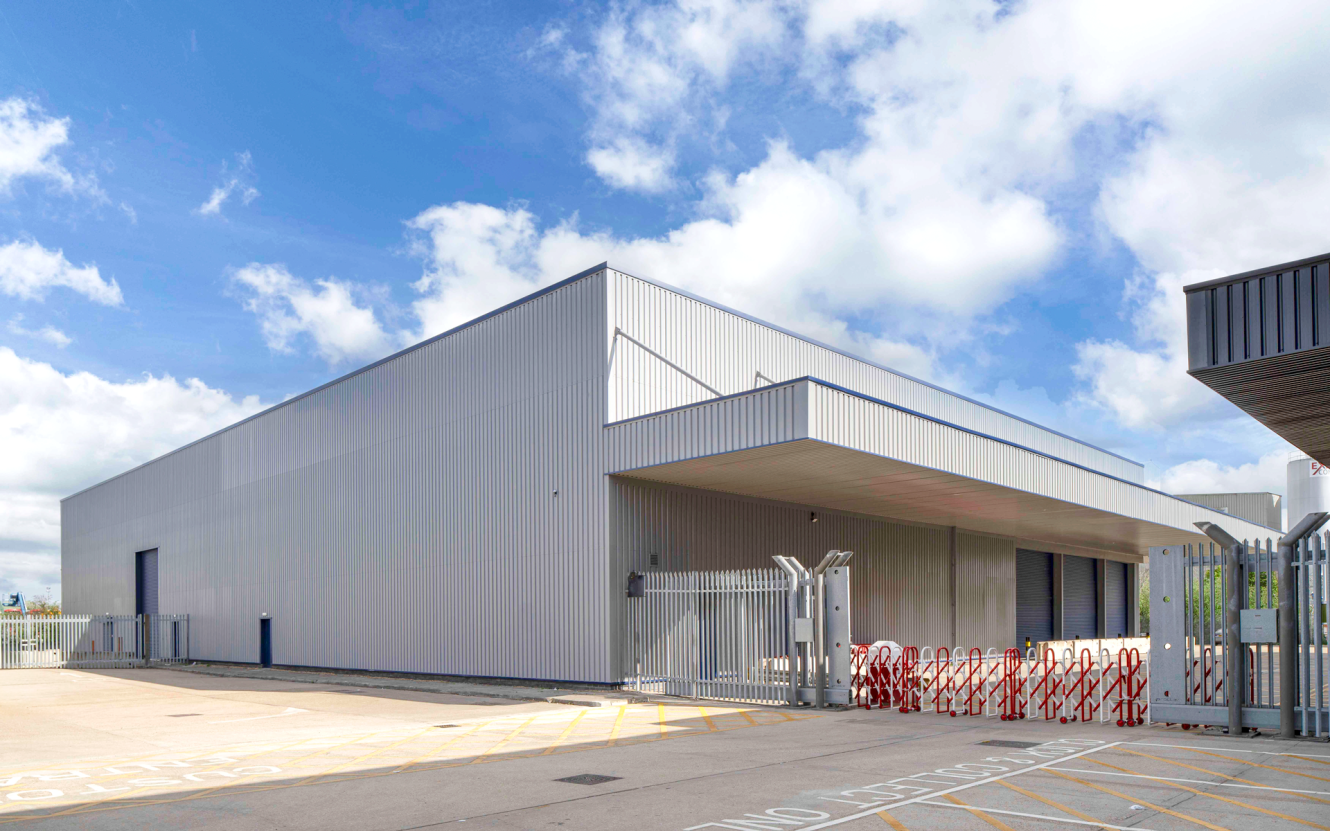 A modern industrial building with a corrugated metal exterior, fenced entrance, bicycle racks, and a clear sky in the background.
