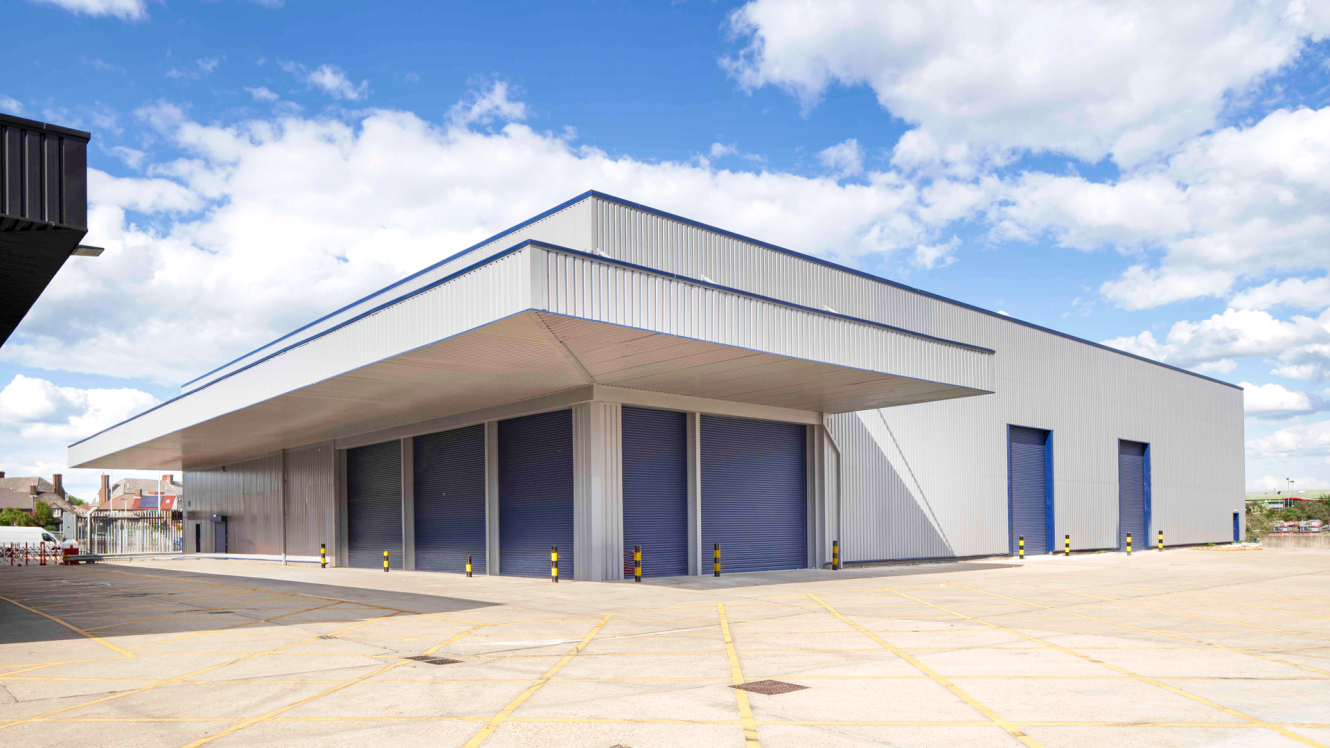 Large industrial warehouse with blue roller shutters, a wide overhang, and a spacious concrete loading area under a partly cloudy sky.
