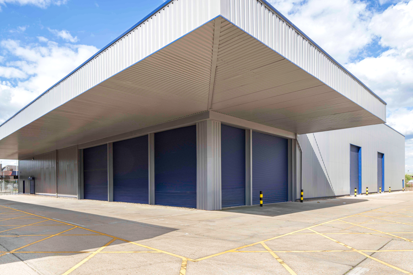 A large modern warehouse featuring blue roller shutter doors and a covered loading bay, bordered by yellow lines on concrete.