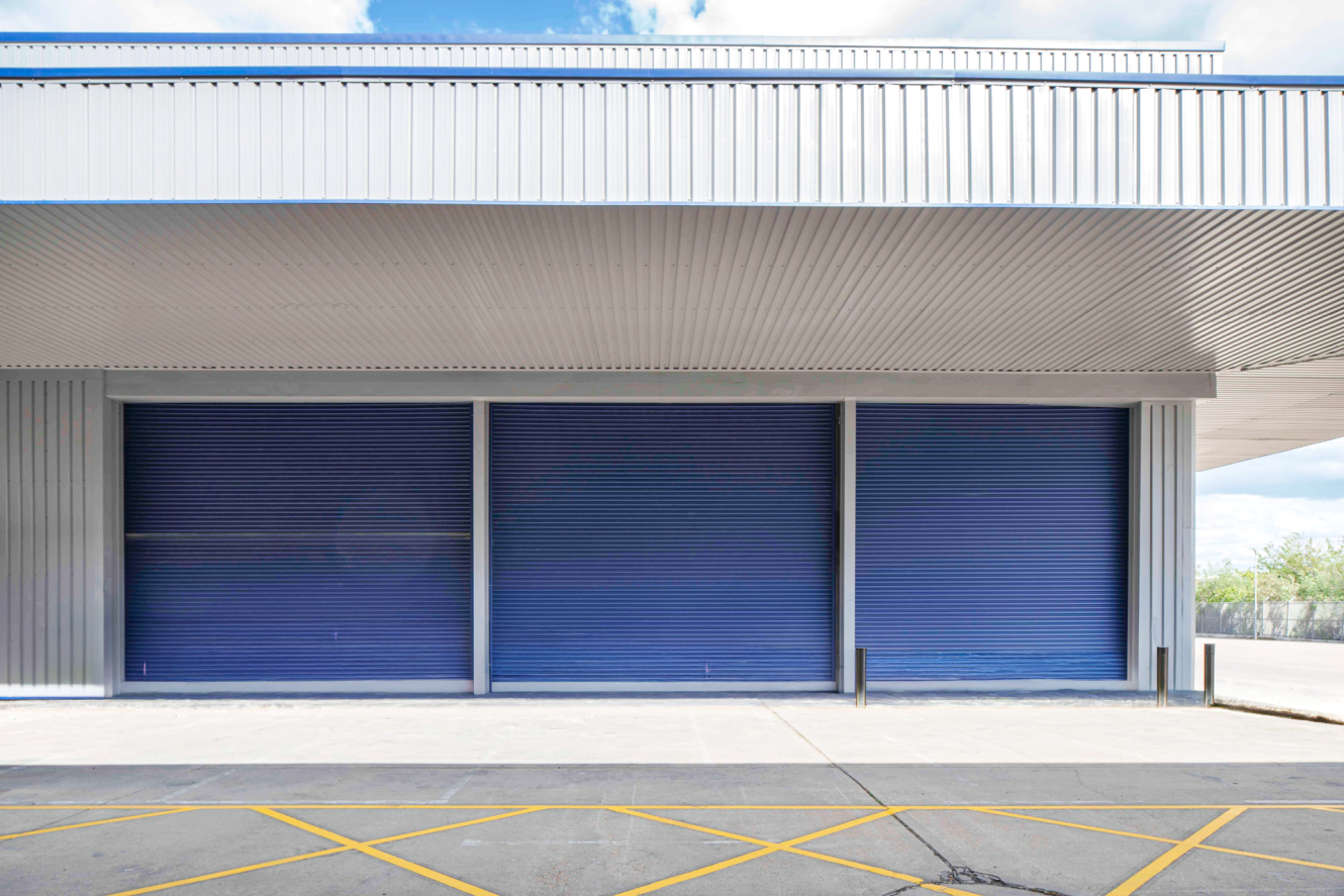 Three large blue roller shutters are closed on an industrial warehouse building, yellow lines marked on the concrete pavement in front.