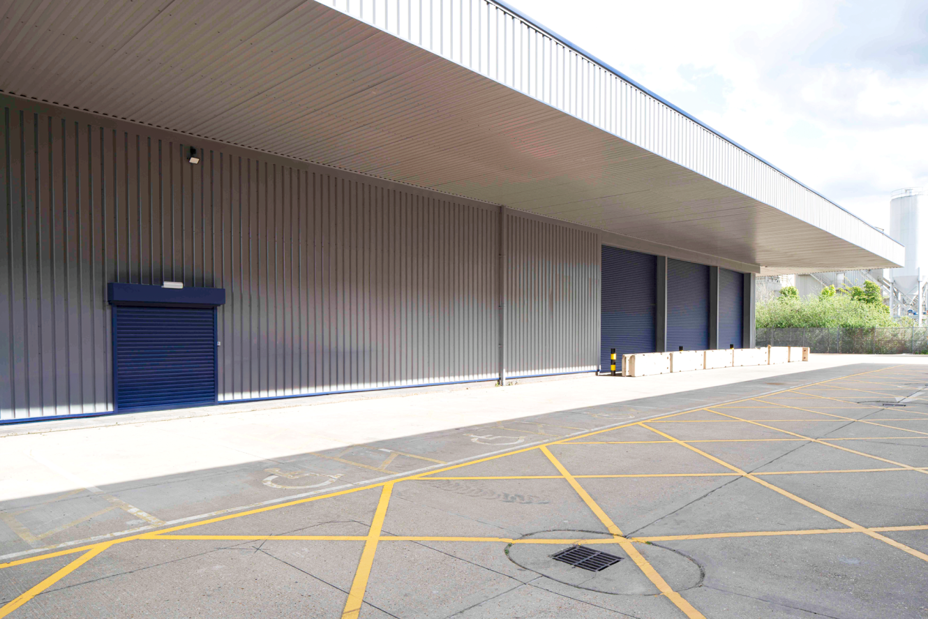 Exterior of a large industrial warehouse with closed blue roller shutters, covered loading bay, and yellow parking lines on concrete.