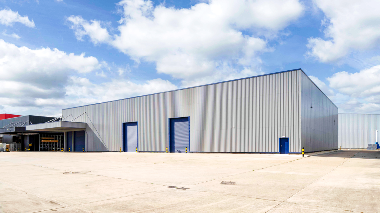 Large, modern industrial warehouse with metal cladding, blue doors, and a wide concrete yard beneath a partly cloudy sky.
