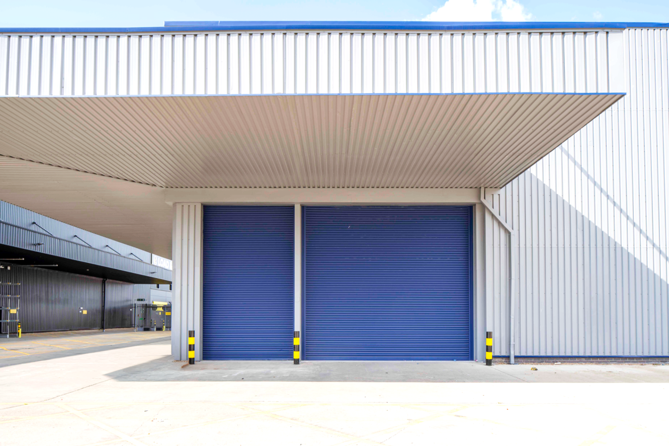 Two large blue roller shutters on a metal industrial warehouse building with a covered loading bay and protective bollards.