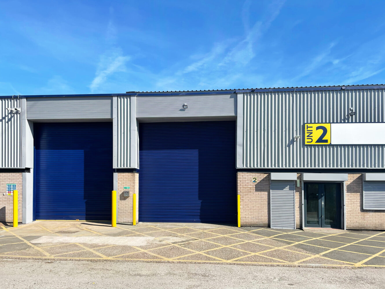 Industrial warehouse with two large blue roller shutters, glass entrance, Unit 2 sign, and yellow markings on the ground outside.