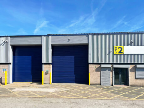Industrial warehouse with two large blue roller shutters, glass entrance, Unit 2 sign, and yellow markings on the ground outside.