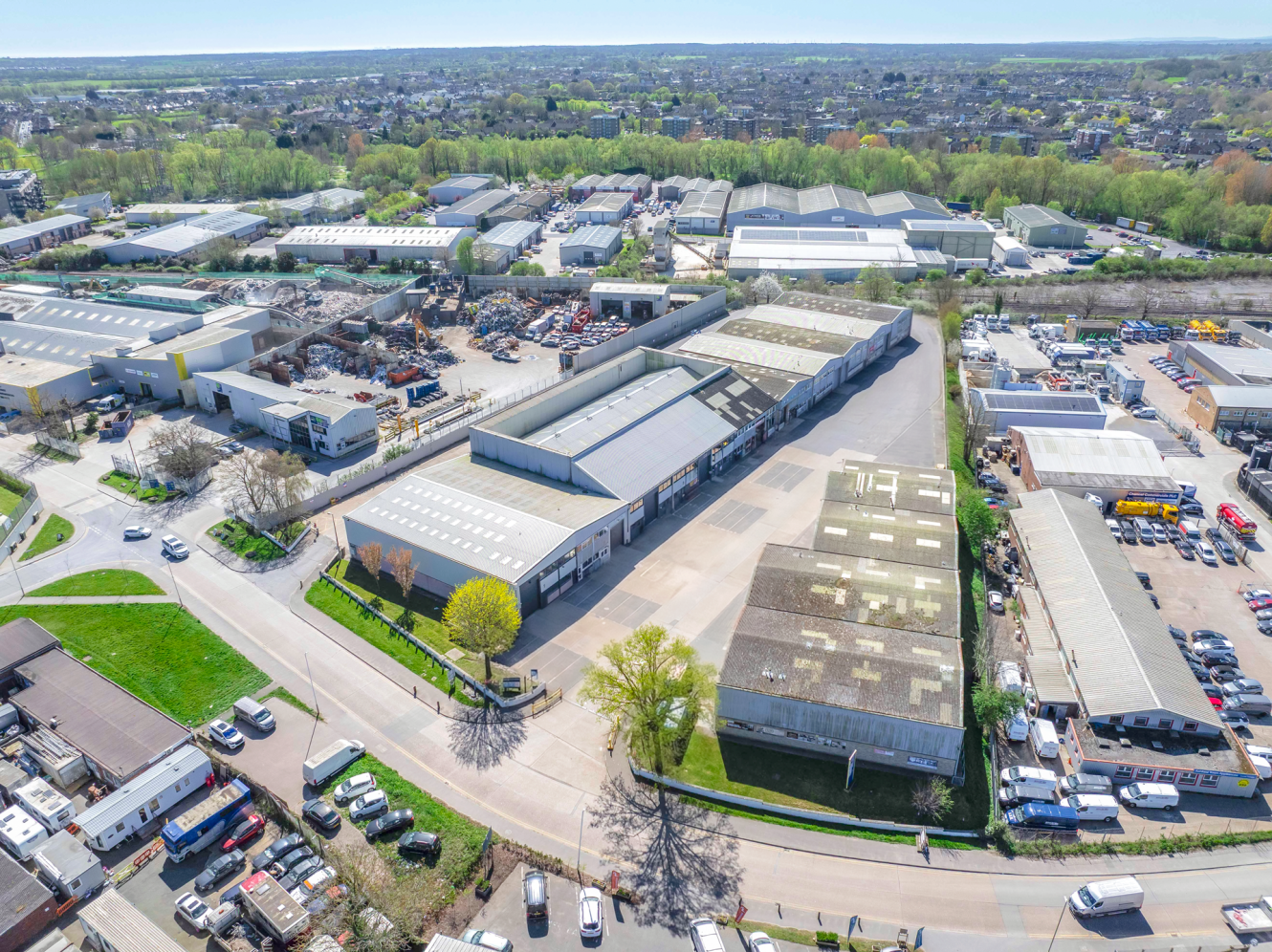 Aerial view of an industrial estate featuring multiple warehouses, roads, parked vehicles, and trees in a suburban setting.