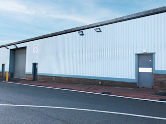 Industrial warehouse exterior with a corrugated metal facade, two doors, overhead lights, and a paved road in front.
