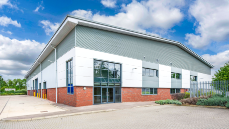 A modern industrial warehouse with large windows, red brick base, and metal cladding on a tarmacked yard beneath a partly cloudy sky.