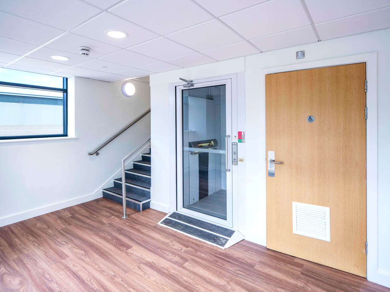 A modern indoor space with wooden floors, a staircase with a metal handrail, a window, a door, and a small lift by the stairs.