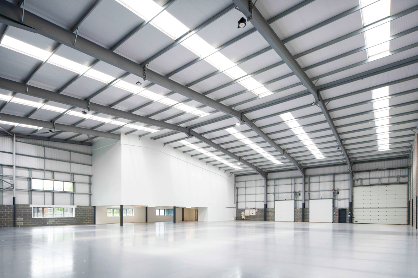 Large, empty industrial warehouse with a high ceiling, skylights, white walls, and wide entrance doors.