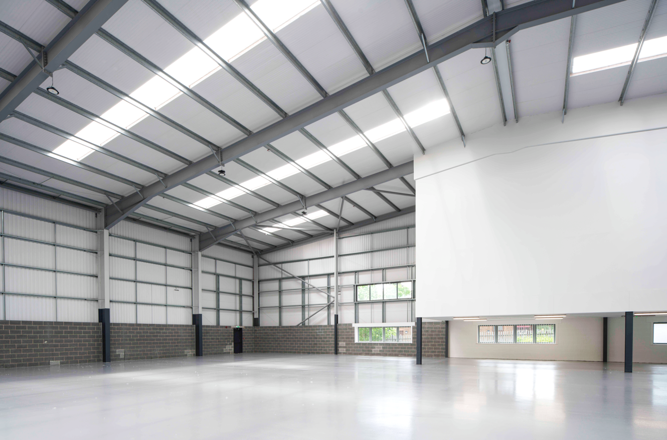 Large, empty industrial warehouse with high ceilings, metal beams, concrete floors, and white walls; natural light streams through windows.