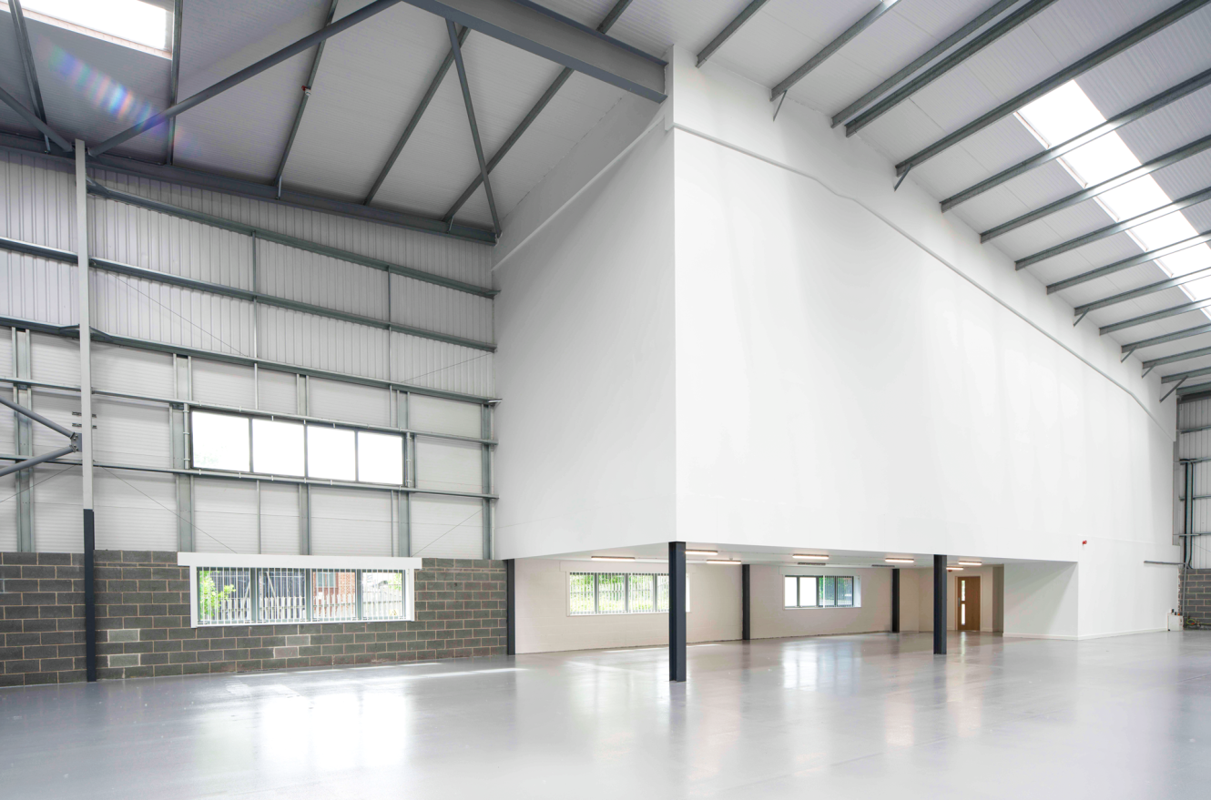Empty modern warehouse interior featuring a high ceiling, grey steel beams, large windows, and a white mezzanine above offices.