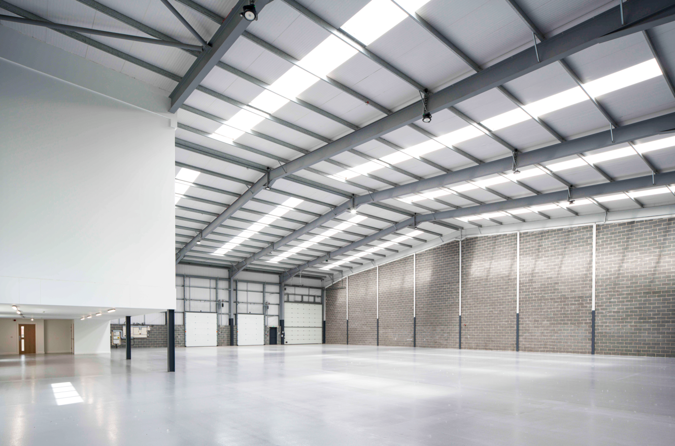 Interior of a spacious, empty warehouse with high ceilings, exposed steel beams, brick walls, polished concrete floor, and loading bay doors.