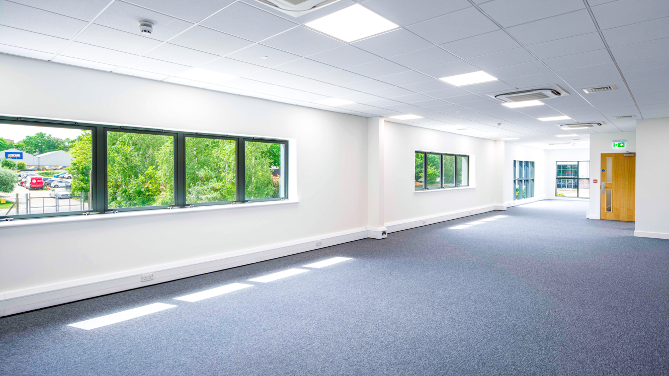 A spacious, empty office room with large windows, white walls, blue carpet, and ceiling lights overlooking greenery and parked cars.