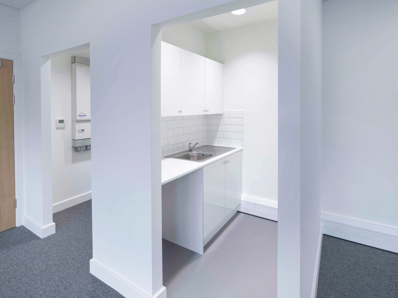 Small modern kitchenette with white cupboards, stainless steel sink, and subway tile splashback in a recessed minimalist space.
