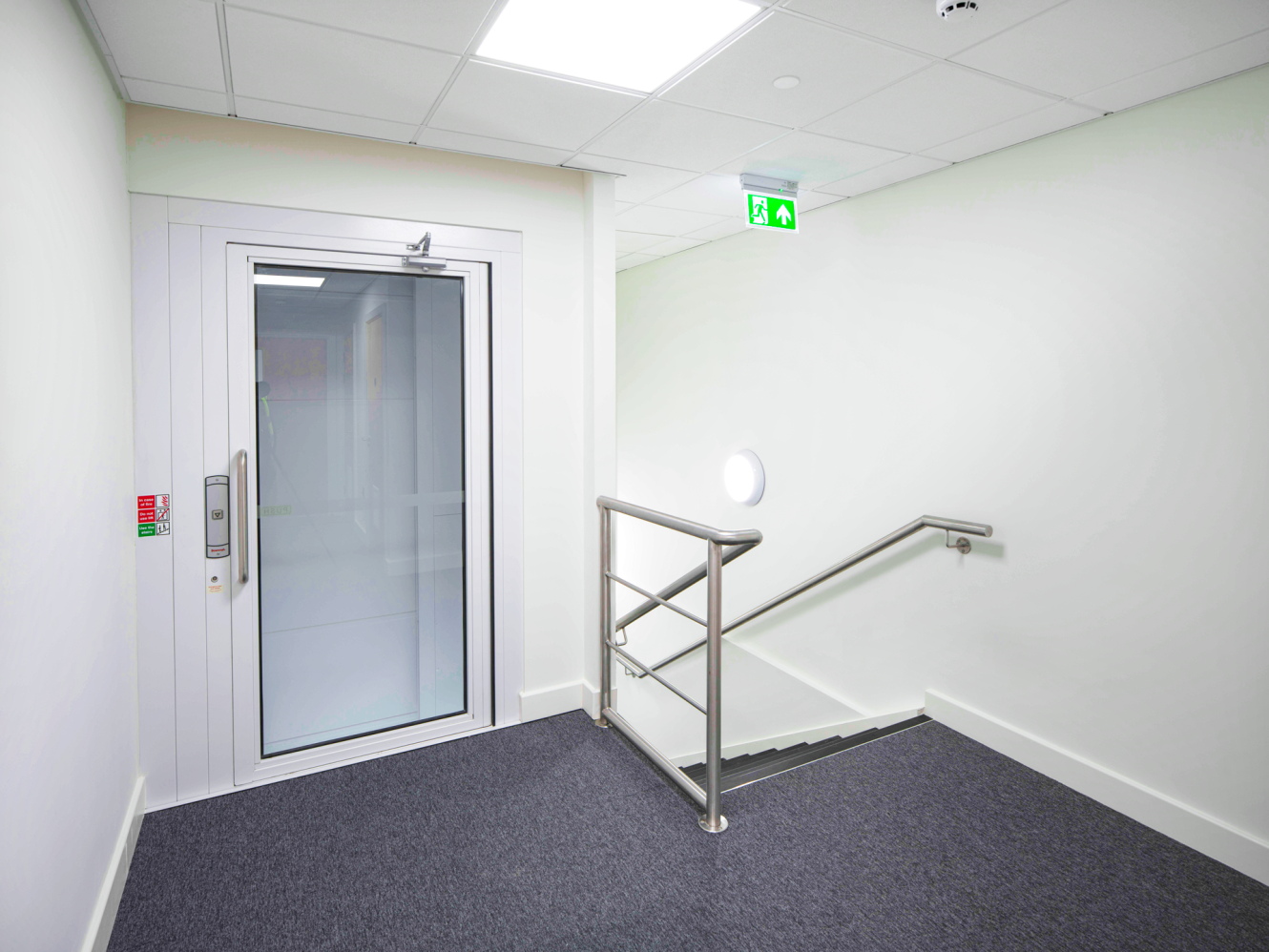 A small lift with glass doors sits beside a stairwell with a metal railing in a well-lit corridor with white walls and grey carpet.