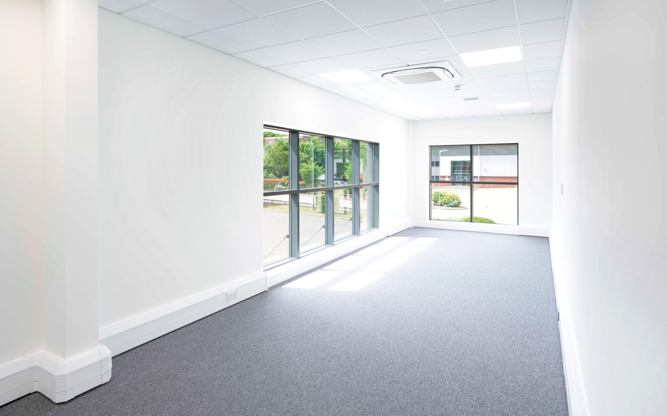 Empty office room with white walls, grey carpet, a large window on the left, and a small window at the end letting in natural light.