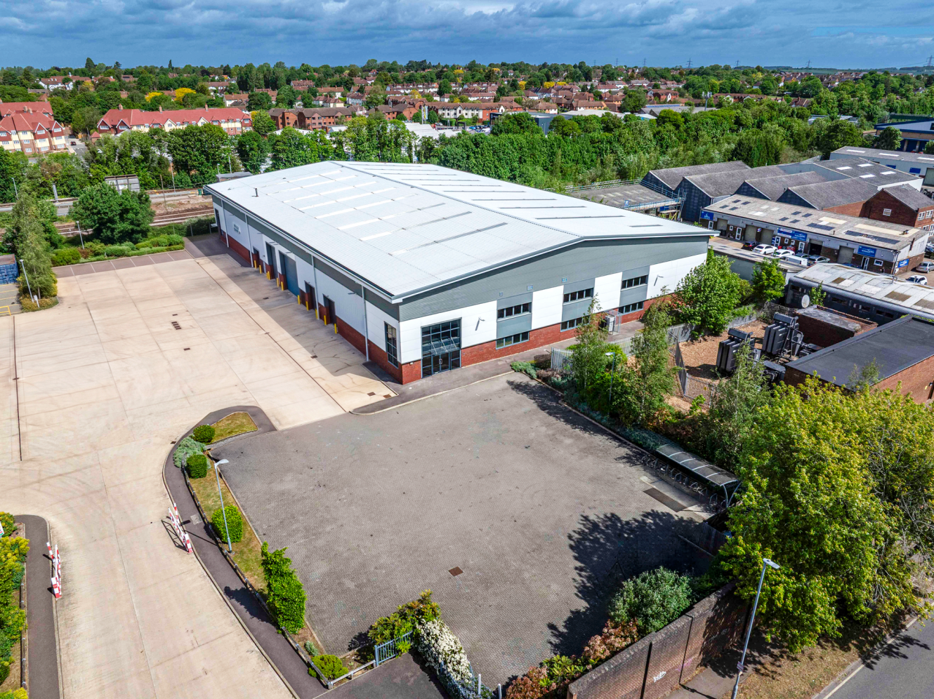 Aerial view of a large industrial warehouse with a white roof, plus paved areas, greenery, and nearby buildings surrounding it.