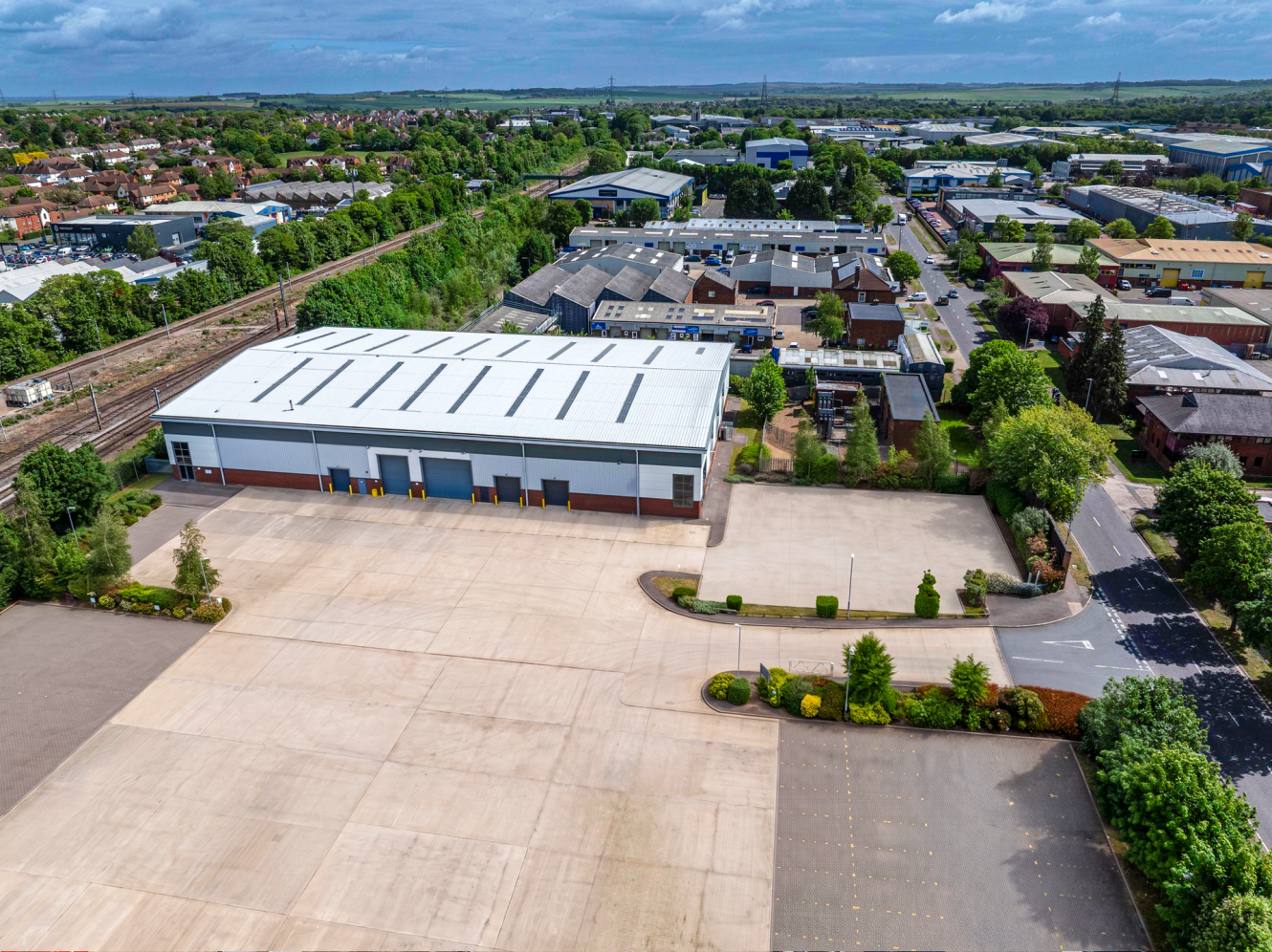 Aerial view of a large industrial warehouse with a wide concrete yard, greenery, and other buildings in an industrial area.