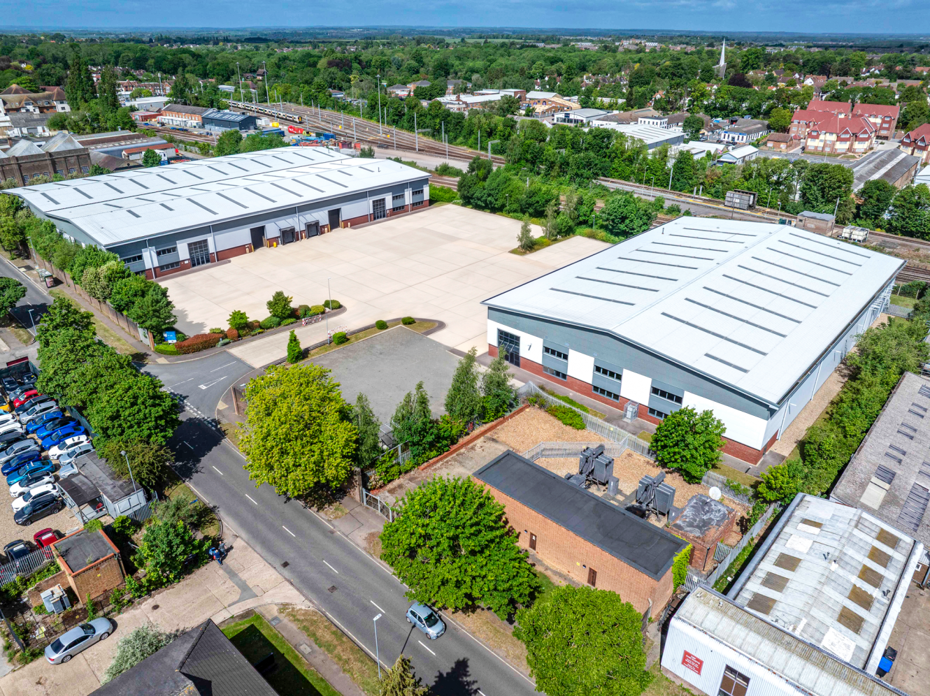 Aerial view of two large industrial warehouses, parking areas, trees, and nearby roads in a suburban setting.