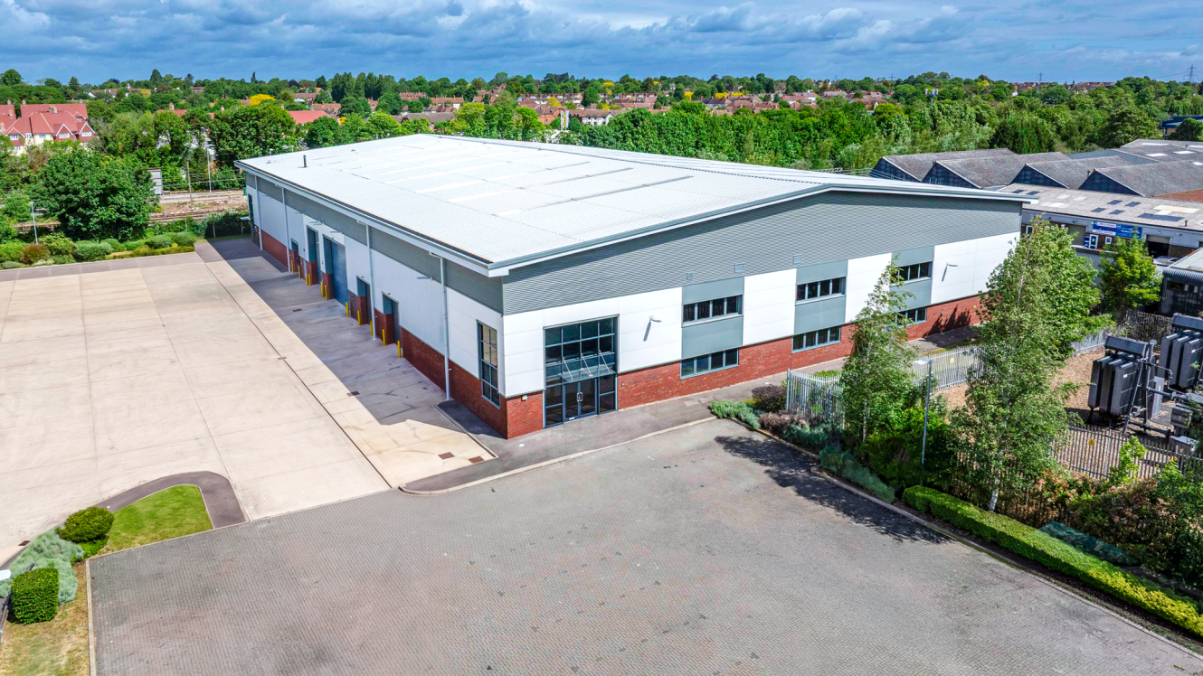 A large, modern warehouse with a white and grey exterior, paved car parks, greenery, and nearby residential and industrial buildings.