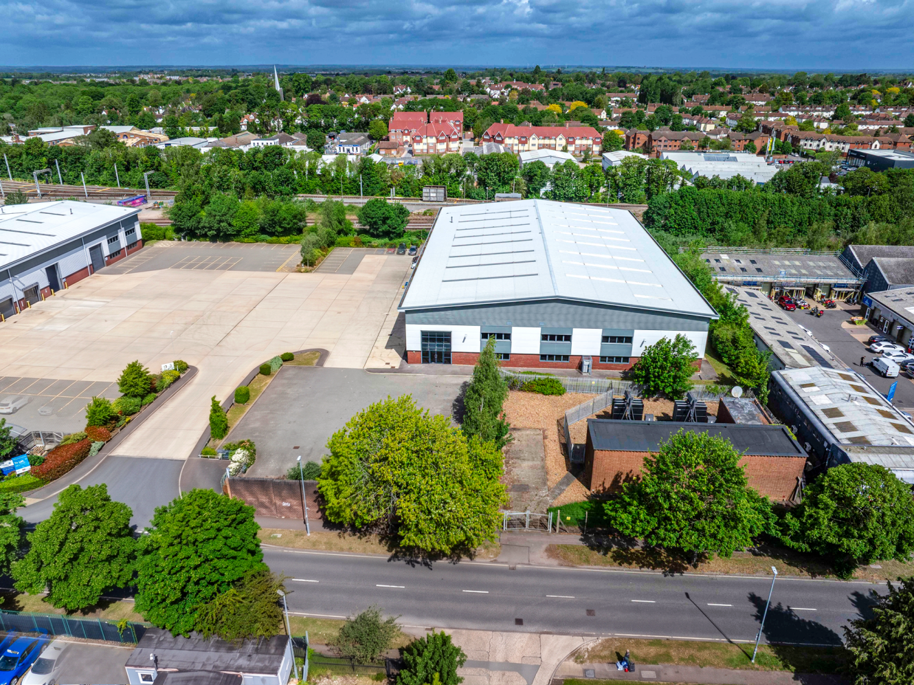 Aerial view of an industrial building with a large empty car park, trees, nearby commercial properties, and homes in the background.
