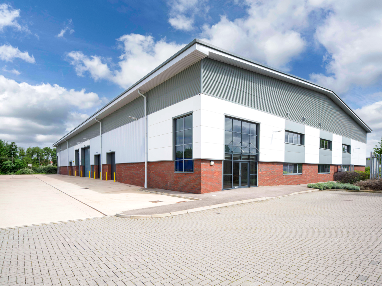 A modern industrial warehouse with large windows and loading bays, surrounded by a paved car park under a partly cloudy sky.