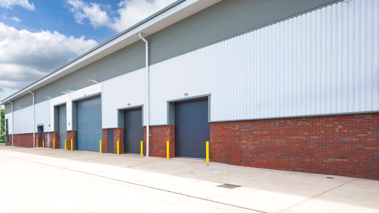 A row of closed warehouse doors with yellow bollards, on a brick and metal building beneath a partly cloudy sky.